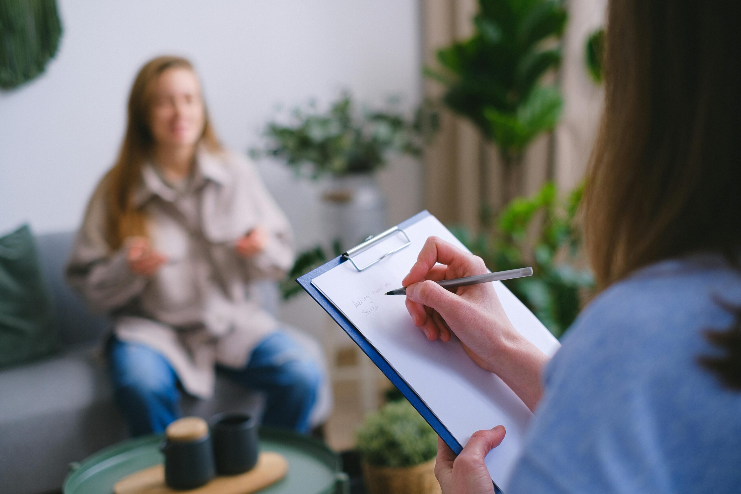 Esileht Unrecognizable professional female psychologist writing on clipboard while sitting against client on blurred background during psychotherapy session in light office